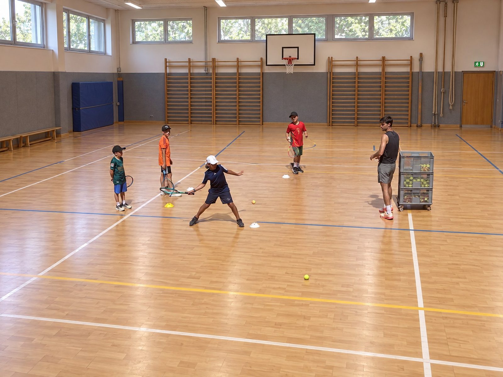 Kind spielt in der Grundschul-Turnhalle eine Vorhand im Stationstraining mit Hütchen, Trainer mit Ballwagen schaut zu